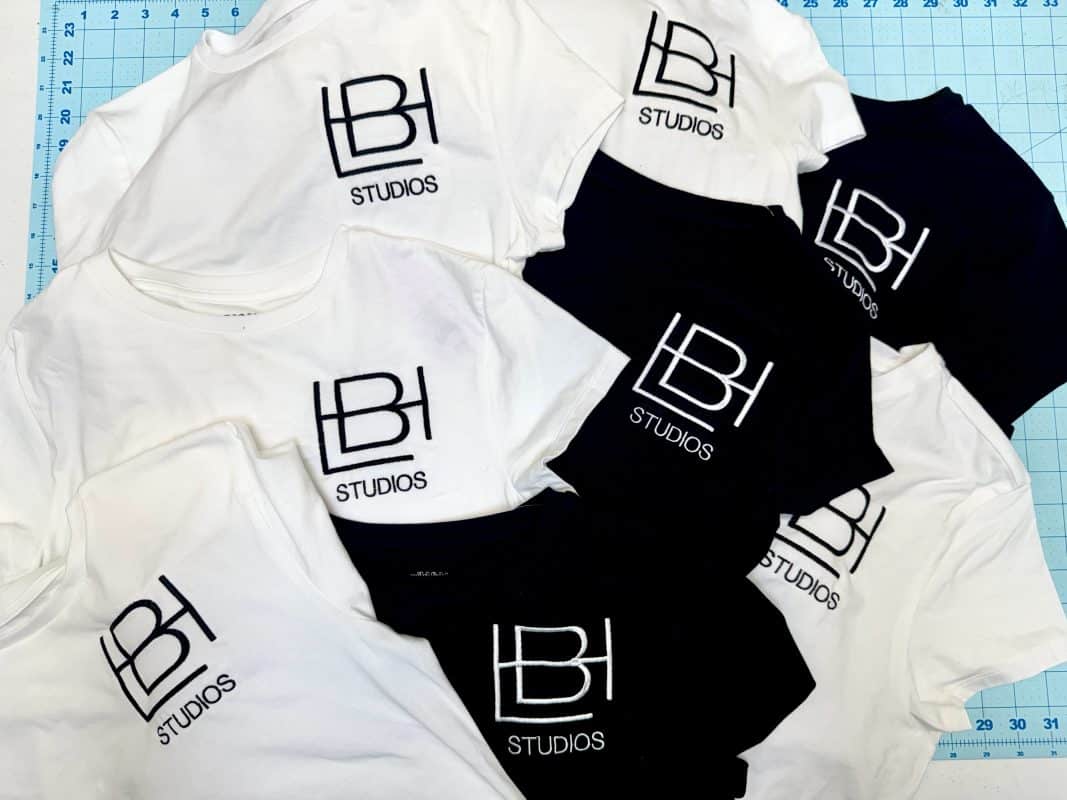 White and black t-shirts with "LBH STUDIOS" logo, arranged on a blue cutting mat, showcasing apparel for Embellishway's product gallery.