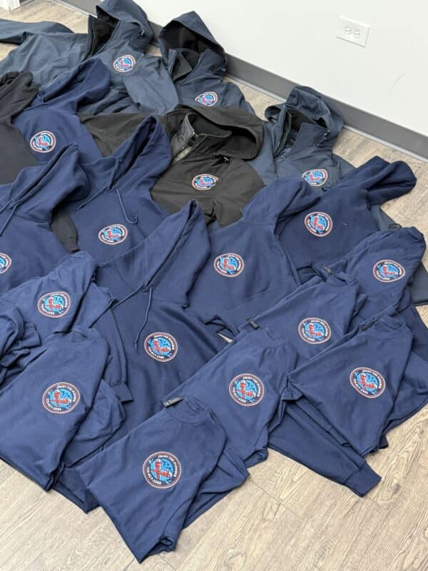 Assorted navy blue hoodies and sweatshirts with logos, displayed on a wooden floor, showcasing products available for sale at Embellishway.