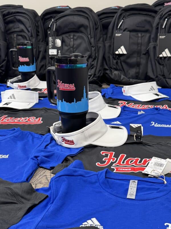 Assorted sports merchandise including blue and black "Flames" T-shirts, white visors, a black travel mug with a skyline design, and black Adidas backpacks, arranged on a table.