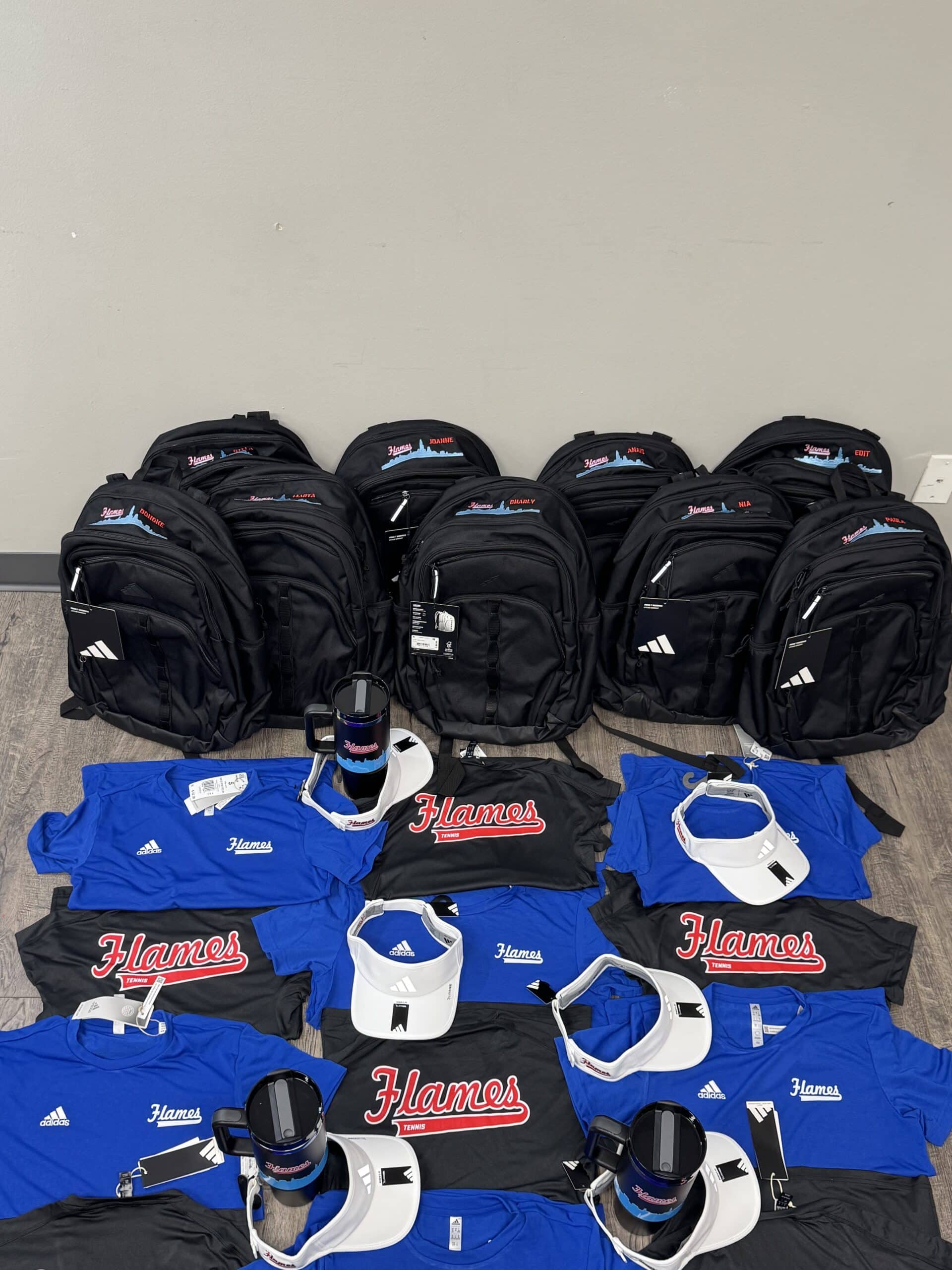 Collection of branded black backpacks, blue and black t-shirts, white visors, and mugs featuring "Flames" logo, showcasing promotional products for EmbellishWay's brand identity and merchandise offerings.