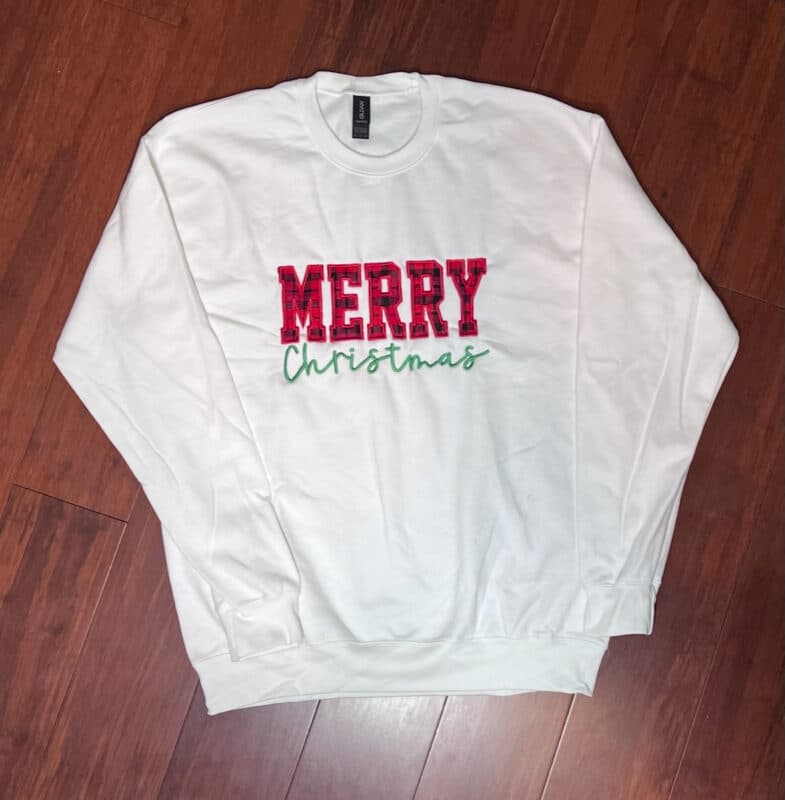 White long-sleeve sweatshirt featuring "MERRY" in red plaid and "Christmas" in green cursive, displayed on wooden floor.