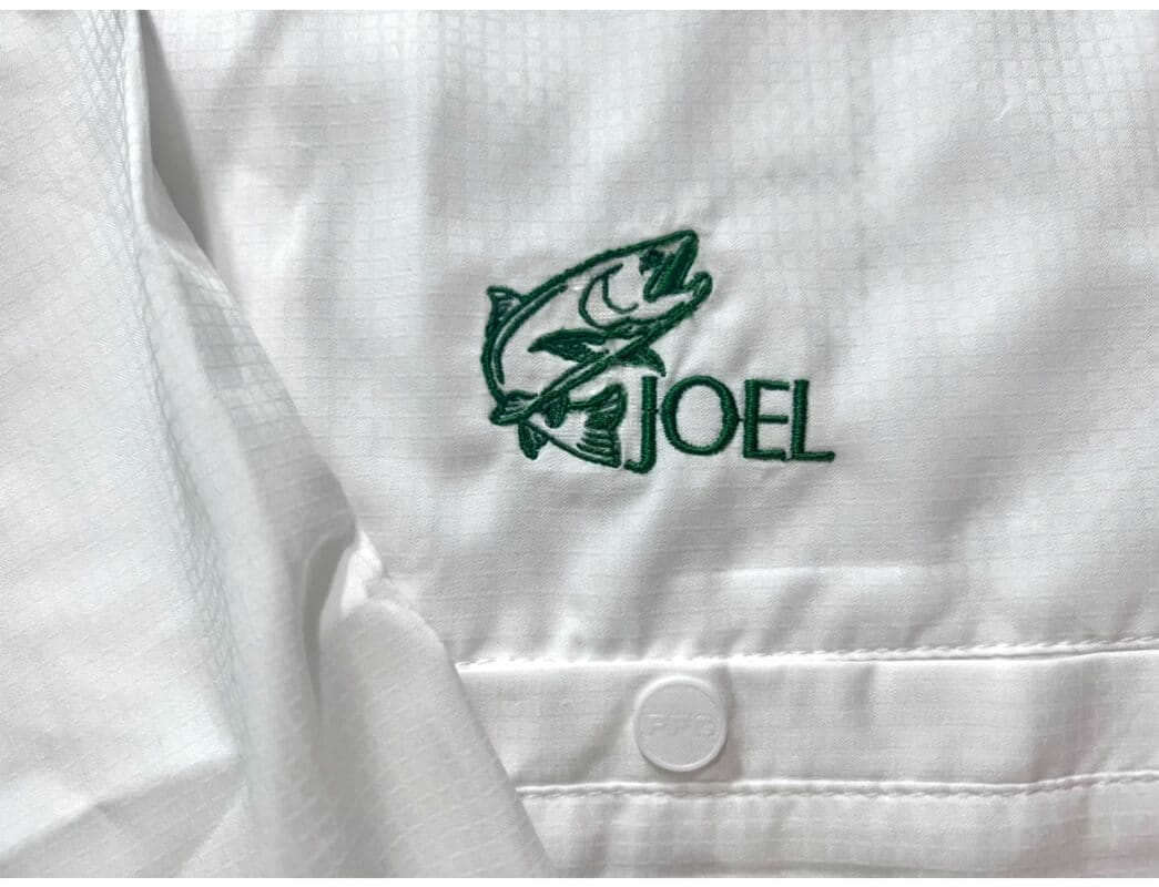 White shirt featuring green embroidered fish logo and the name "JOEL," suitable for branding in outdoor or fishing apparel.
