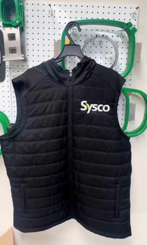 Black sleeveless vest with Sysco logo displayed on a pegboard background featuring green equipment.