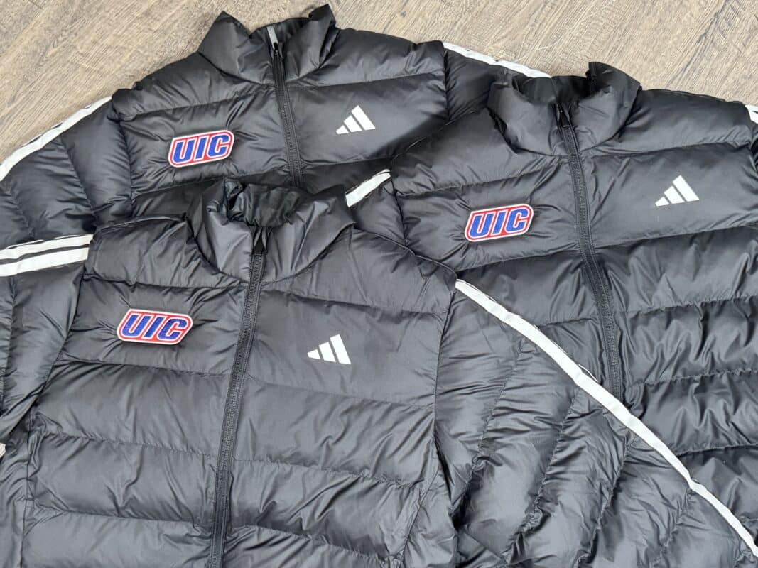 Black puffer jackets featuring UIC logo and Adidas branding, displayed on a wooden surface.