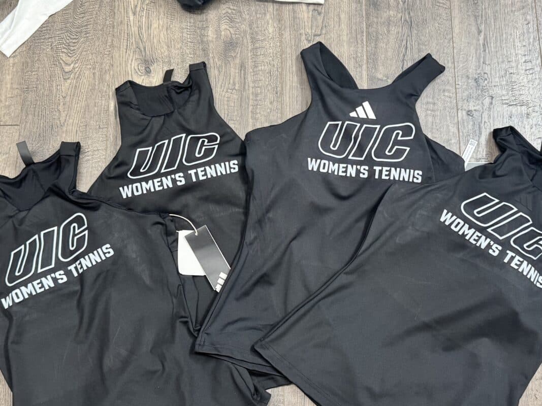 Black athletic tank tops with "UIC WOMEN'S TENNIS" printed in white, displayed on a wooden surface, showcasing the University of Illinois at Chicago branding for women's tennis apparel.