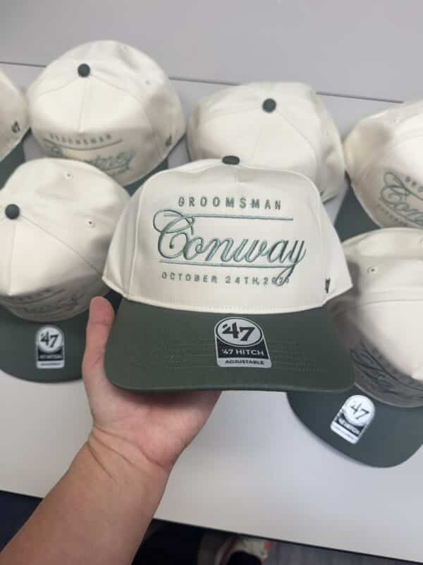 Groomsman hat with "Conway" and date "October 24th 2023," held by a hand, surrounded by similar hats, showcasing personalized wedding accessories.
