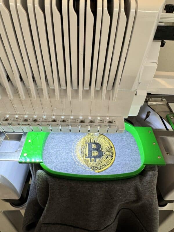 Embroidery machine stitching a Bitcoin logo on fabric, showcasing custom apparel creation for Embellishway's product gallery.