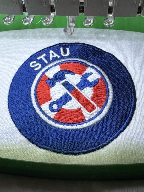 Embroidery machine stitching a circular logo with the text "STAU" and crossed tools, showcasing craftsmanship in product design for Embellishway's gallery.