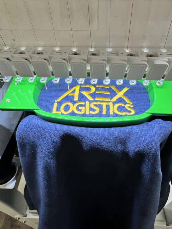 Embroidered logo of "AREX LOGISTICS" on blue fabric, displayed on an embroidery machine.