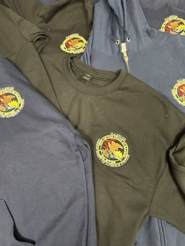 Assorted sweatshirts and hoodies in dark colors featuring the Illinois state emblem, showcasing products available for purchase at Embellishway.