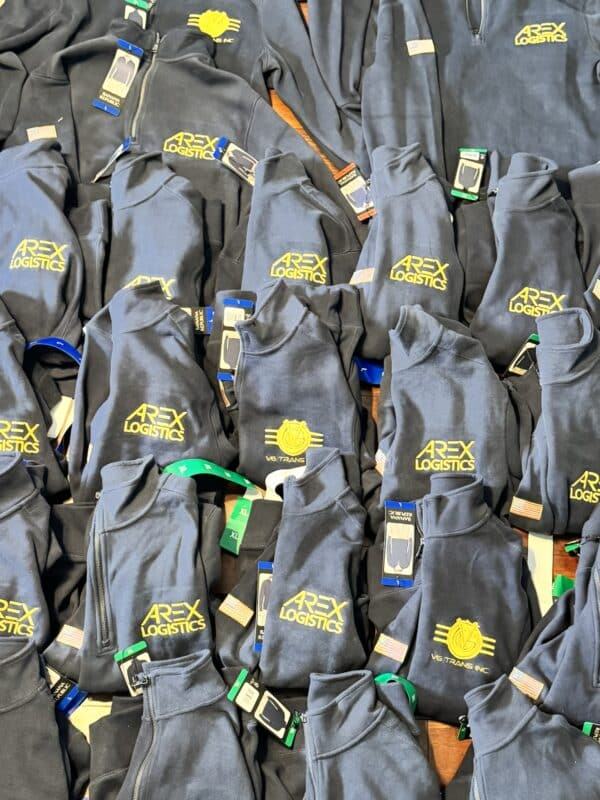 Piles of navy blue fleece jackets with yellow "AREX LOGISTICS" logo and tags, showcasing apparel for promotional or work purposes.